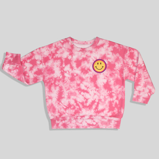 A sustainable, fun, and comfortable pink and white tie-dye crewneck sweatshirt for kids featuring a yellow smiley face graphic with a purple and pink outline on the left chest, displayed against a solid gray background.