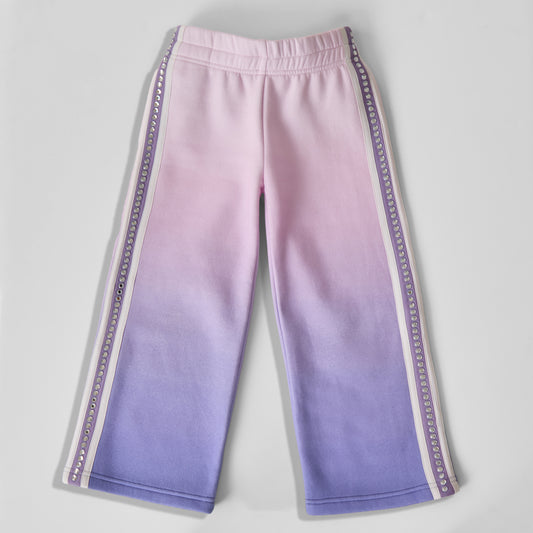 A pair of eco-friendly, fun, and comfortable wide-leg sweatpants featuring a pink-to-purple ombré gradient pattern, with decorative purple and white side stripes and a line of recycled rhinestones down the legs, laid flat on a light gray background.