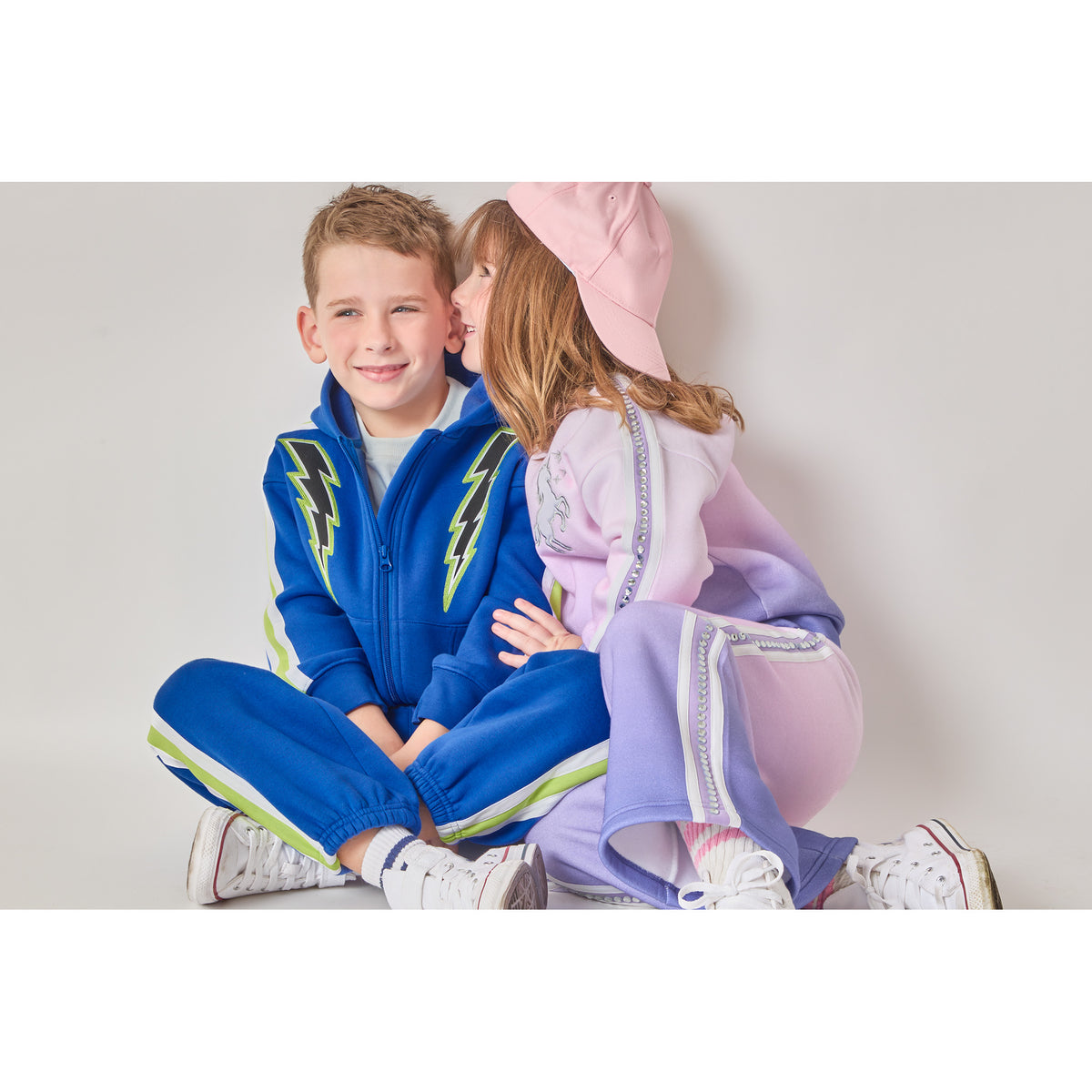 Two children sitting together in eco-friendly outfits. The boy is wearing a blue zip-up hoodie with lightning bolts, and the girl is wearing a sweatshirt and sweatpants with a pink-to-purple ombré pattern, and a row of recycled rhinestones down the sides.
