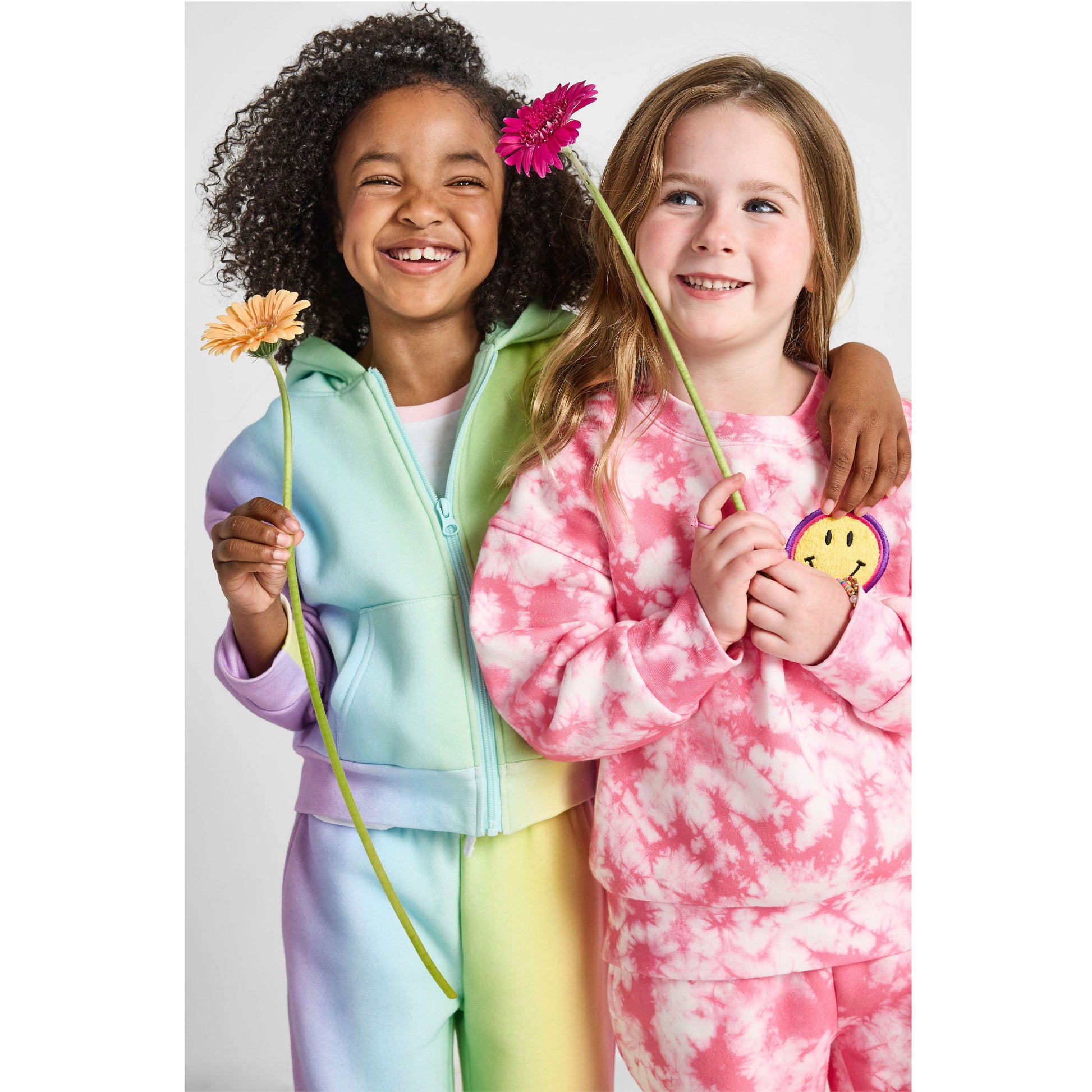 girls holding daisies and wearing our sustainable rainbow wide-leg jogger set and smiley tie-dye jogger set