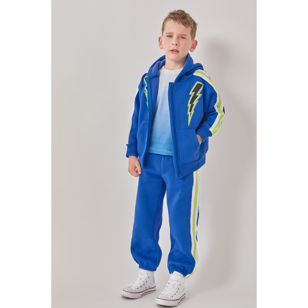 A boy in a sustainable blue lightning bolt sweatshirt, paired with a white-to-blue ombré t-shirt, and matching blue joggers with white and green side stripes.