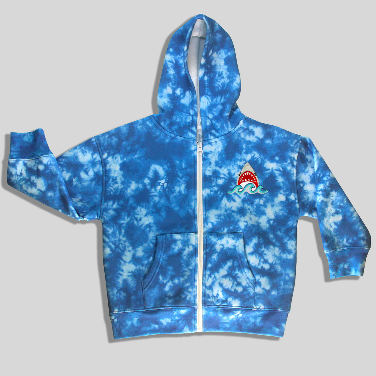 A sustainable, fun, and comfortable blue and white tie-dye zip-up hoodie for children with an embroidered shark graphic on the left chest, displayed against a solid gray background.