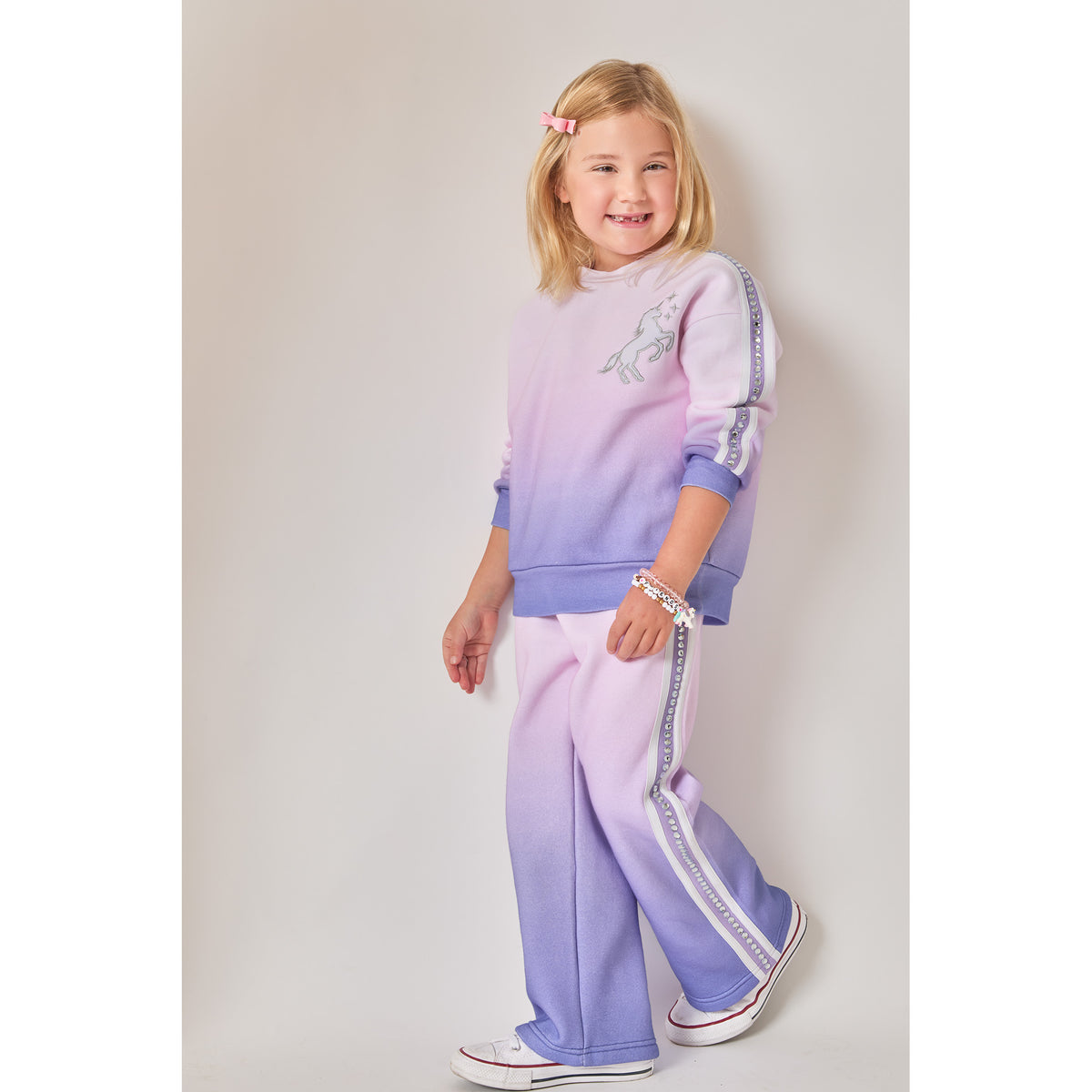 A girl wearing a fun, eco-friendly matching two-piece sweat set with a purple and pink ombre gradient. The set includes a sweatshirt with a unicorn graphic and matching wide-leg joggers. Both have decorative side stripes and recycled rhinestones down the arms and legs.