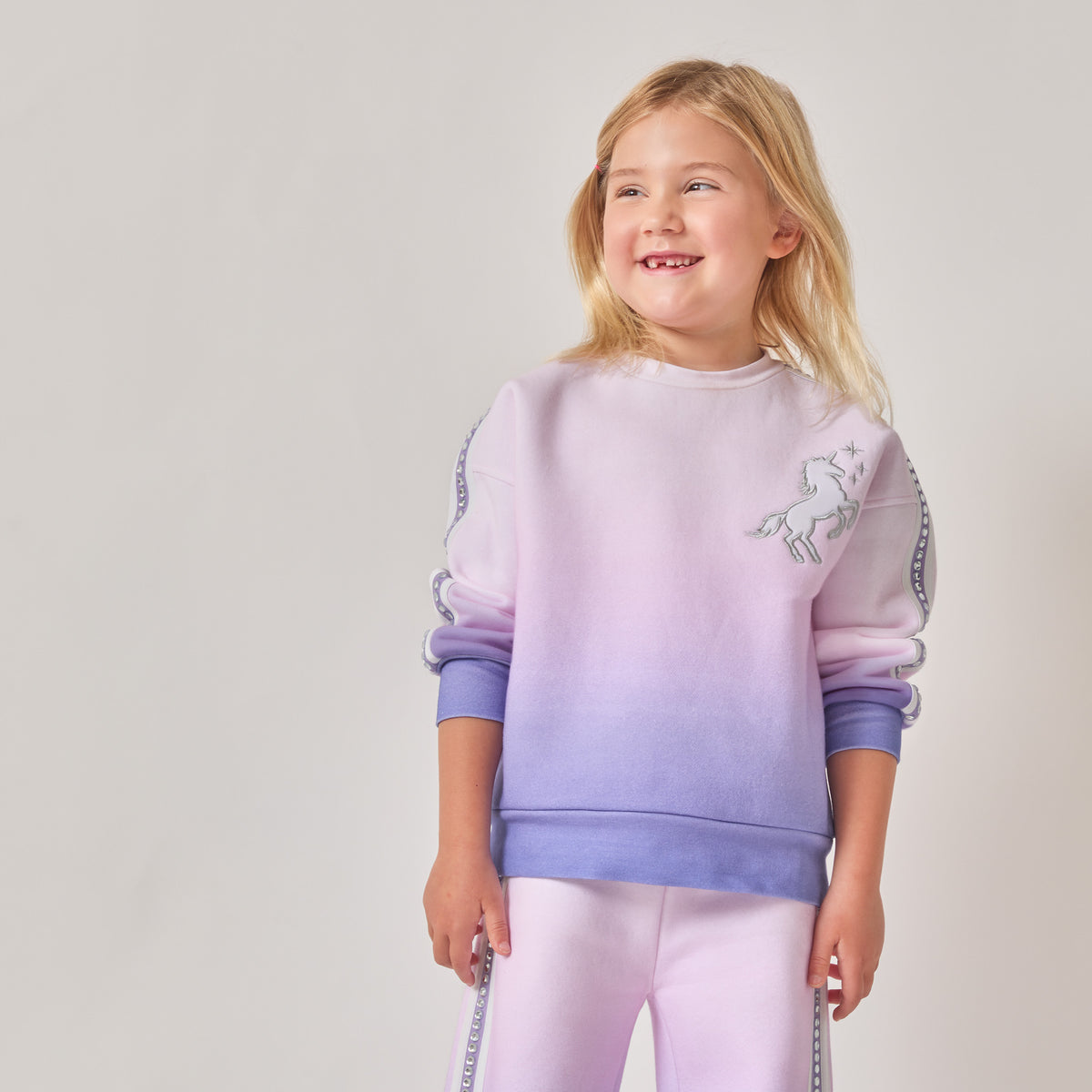 A young girl with blonde hair, wearing a sustainable purple and pink ombré sweatshirt with a white unicorn graphic on the chest, paired with matching ombré pants.
