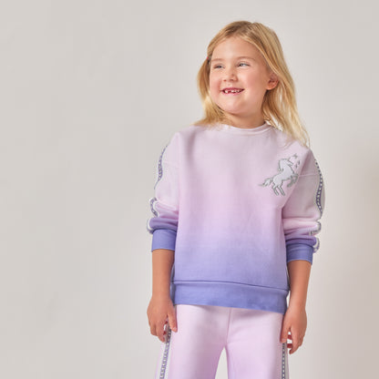 A young girl with blonde hair, wearing a sustainable purple and pink ombré sweatshirt with a white unicorn graphic on the chest, paired with matching ombré pants.