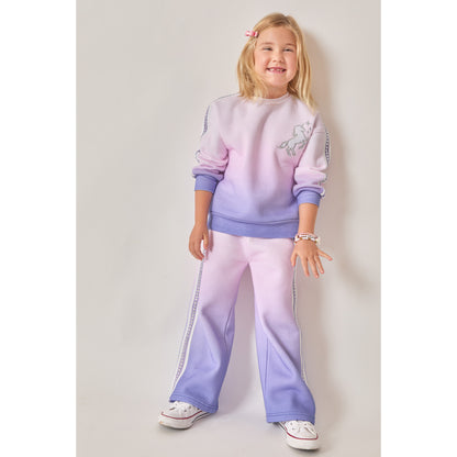 A girl wearing a matching eco-friendly two-piece lounge set with a purple-to-pink ombré gradient. The sweatshirt features a white unicorn graphic on the chest, and the wide-leg joggers have white stripes down the side.  Both have silver recycled rhinestones down the sides.