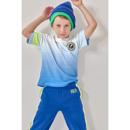 A boy in an eco-friendly organic cotton blue ombré t-shirt with green sleeve stripes and a circular lightning chest patch, paired with matching blue sweatpants featuring green and white side stripes and 'RBLM' embroidered letters near the waistband.