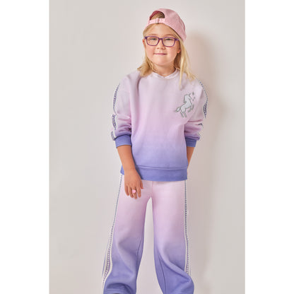 A girl wearing a sustainable purple and pink ombré unicorn sweatshirt with matching wide-leg sweatpants. Both have recycled rhinestones down the arms and legs.