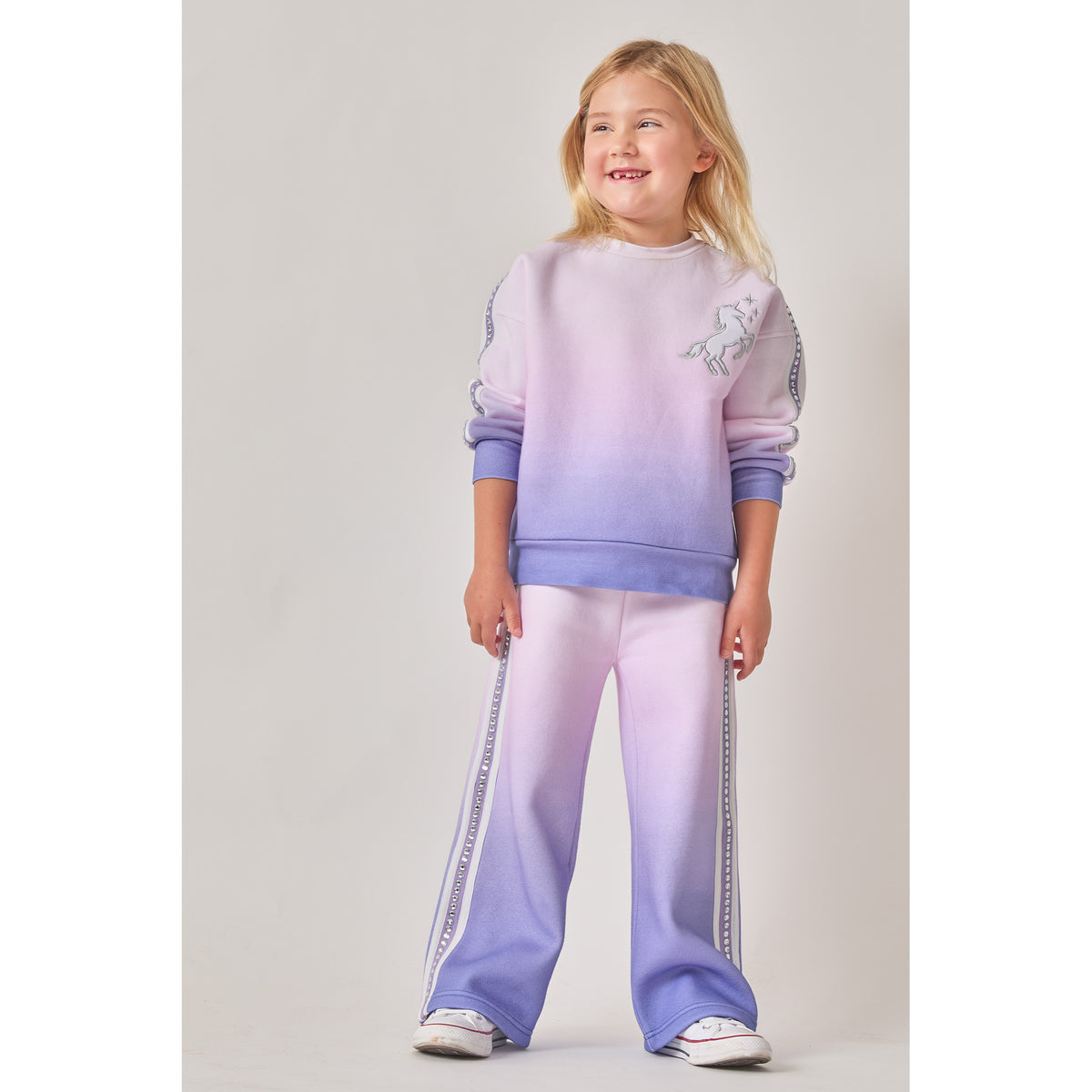 A girl wearing an eco-friendly matching two-piece sweat set with a purple-to-pink ombre gradient pattern. The sweatshirt features a white unicorn graphic on the chest. Both the sweatshirt and wide-leg joggers have purple and white fabric stripes, bedazzled with silver recycled rhinestones, down the sides.
