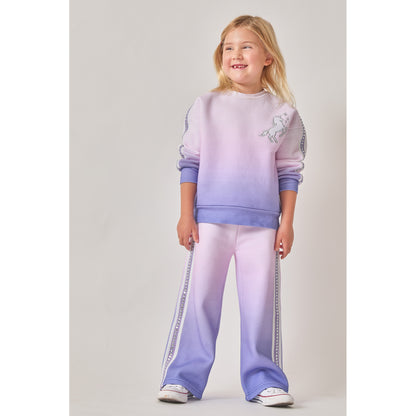 A girl wearing an eco-friendly matching two-piece sweat set with a purple-to-pink ombre gradient pattern. The sweatshirt features a white unicorn graphic on the chest. Both the sweatshirt and wide-leg joggers have purple and white fabric stripes, bedazzled with silver recycled rhinestones, down the sides.
