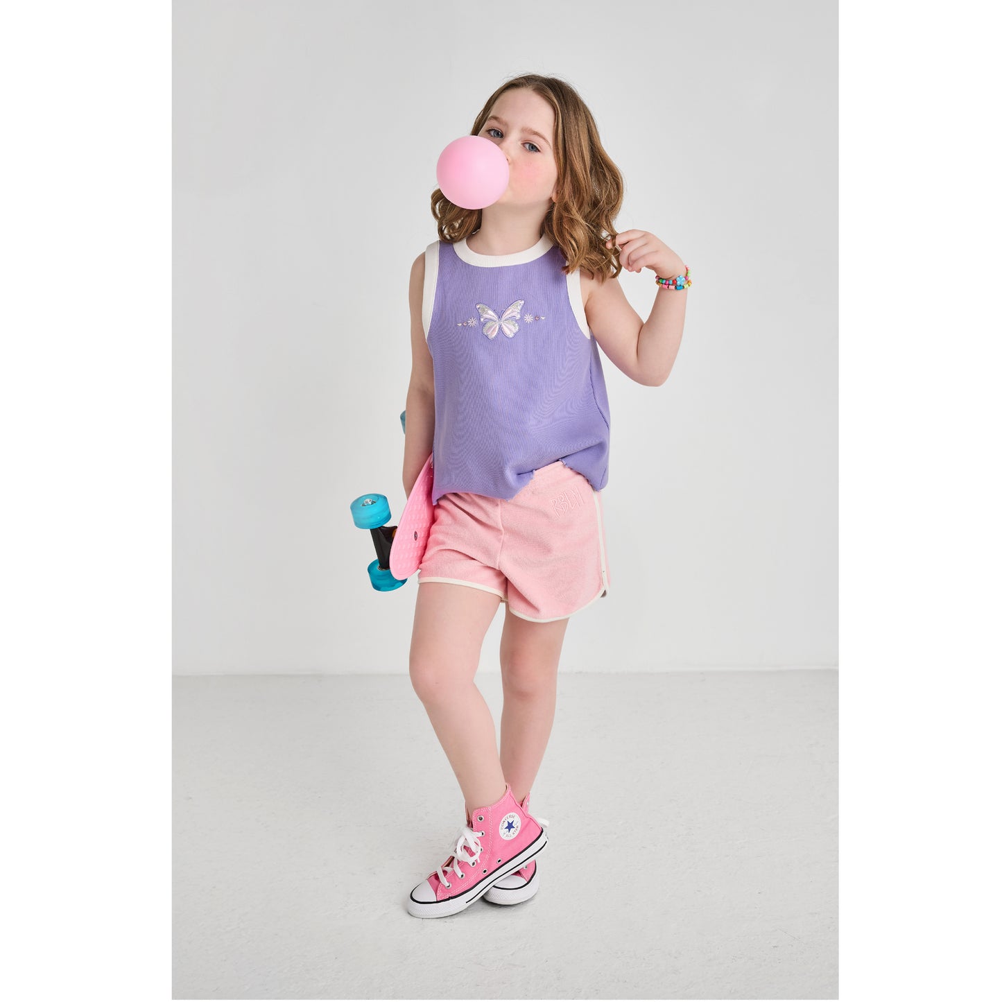 A skateboarding girl wearing a sustainable, purple butterfly tank top and retro pink shorts.