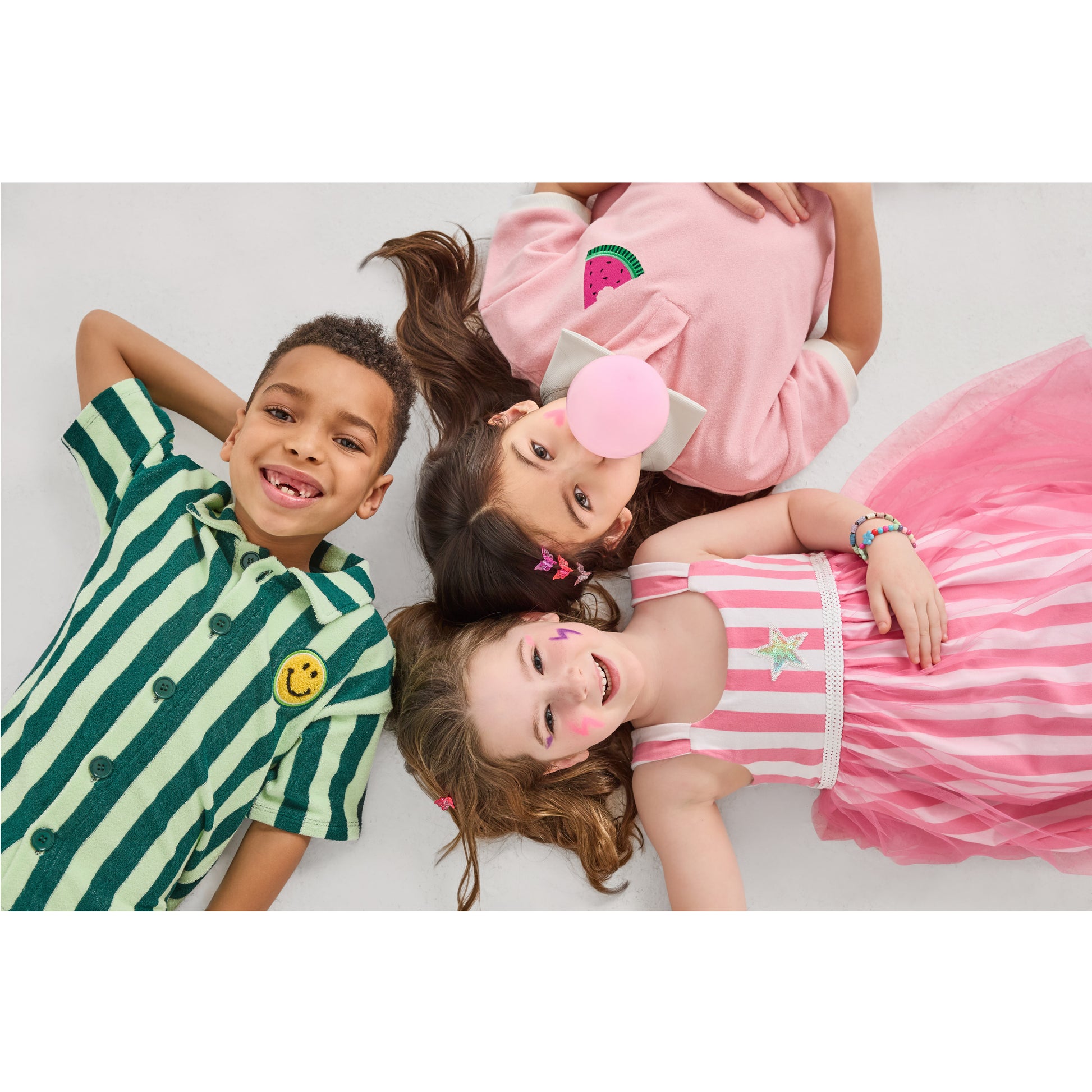 Three children lying on a white surface, wearing sustainable, fun, colorful outfits.