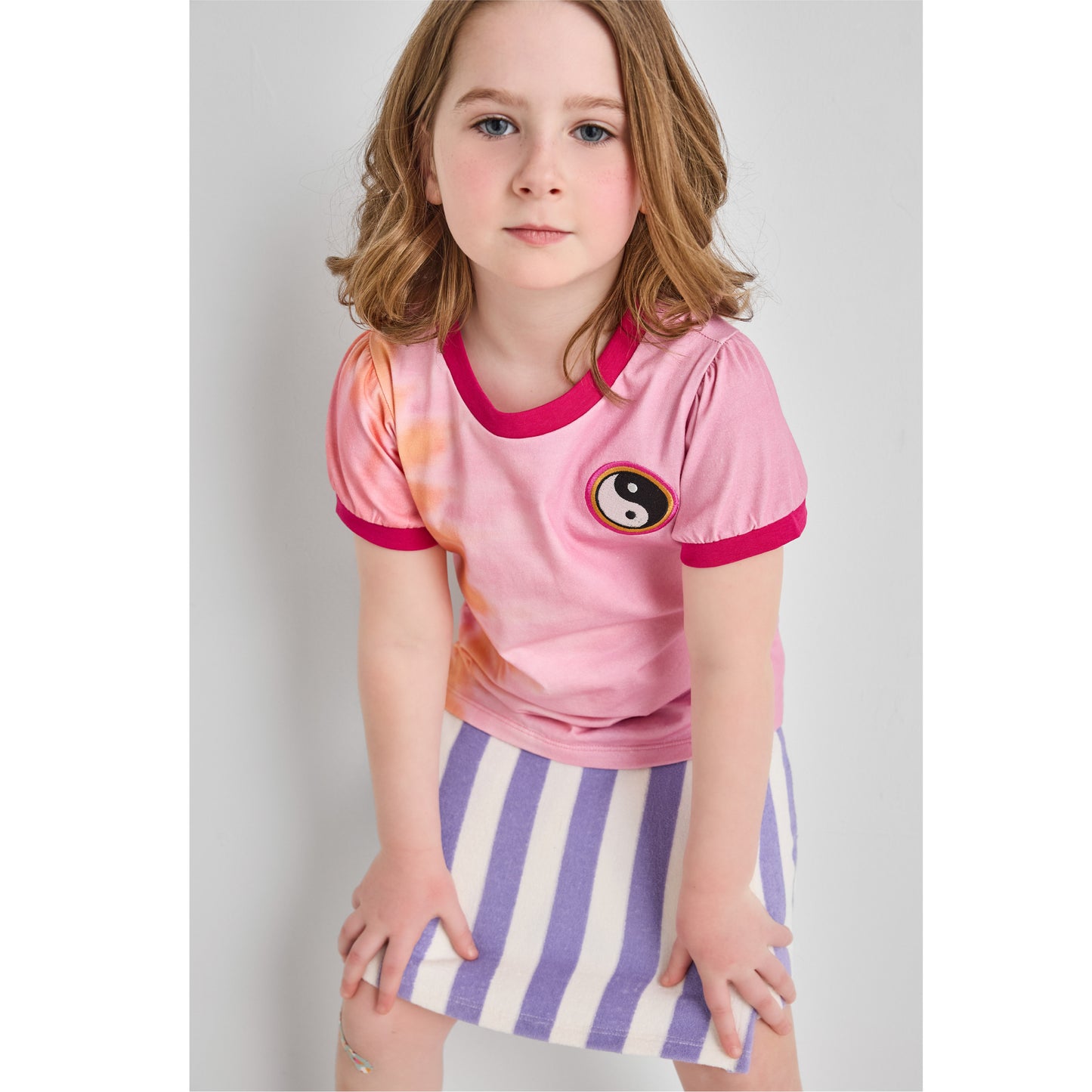 Girl wearing a sustainable, pink retro tie-dye t-shirt with a ying-yang patch and a purple striped skort 