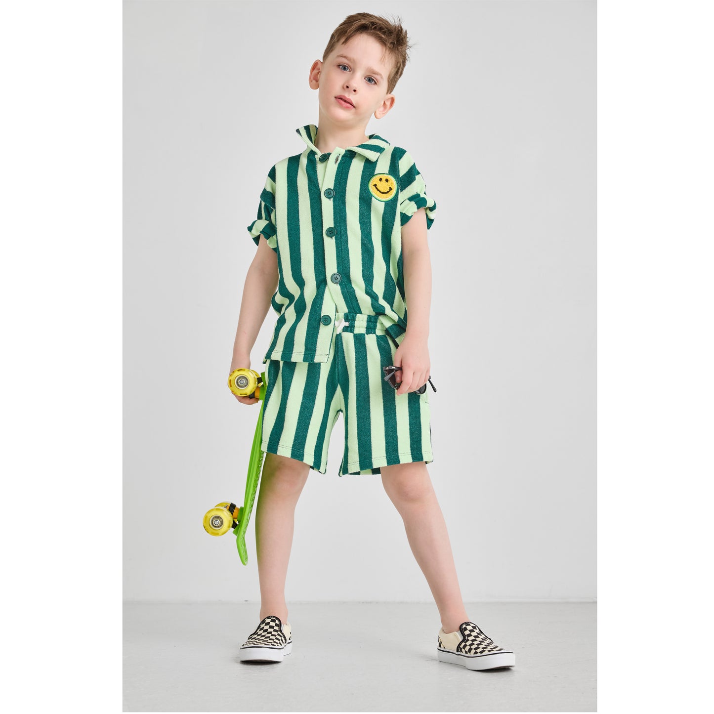 Boy wearing a sustainable, bright green striped set with a smiley face design, holding a skateboard