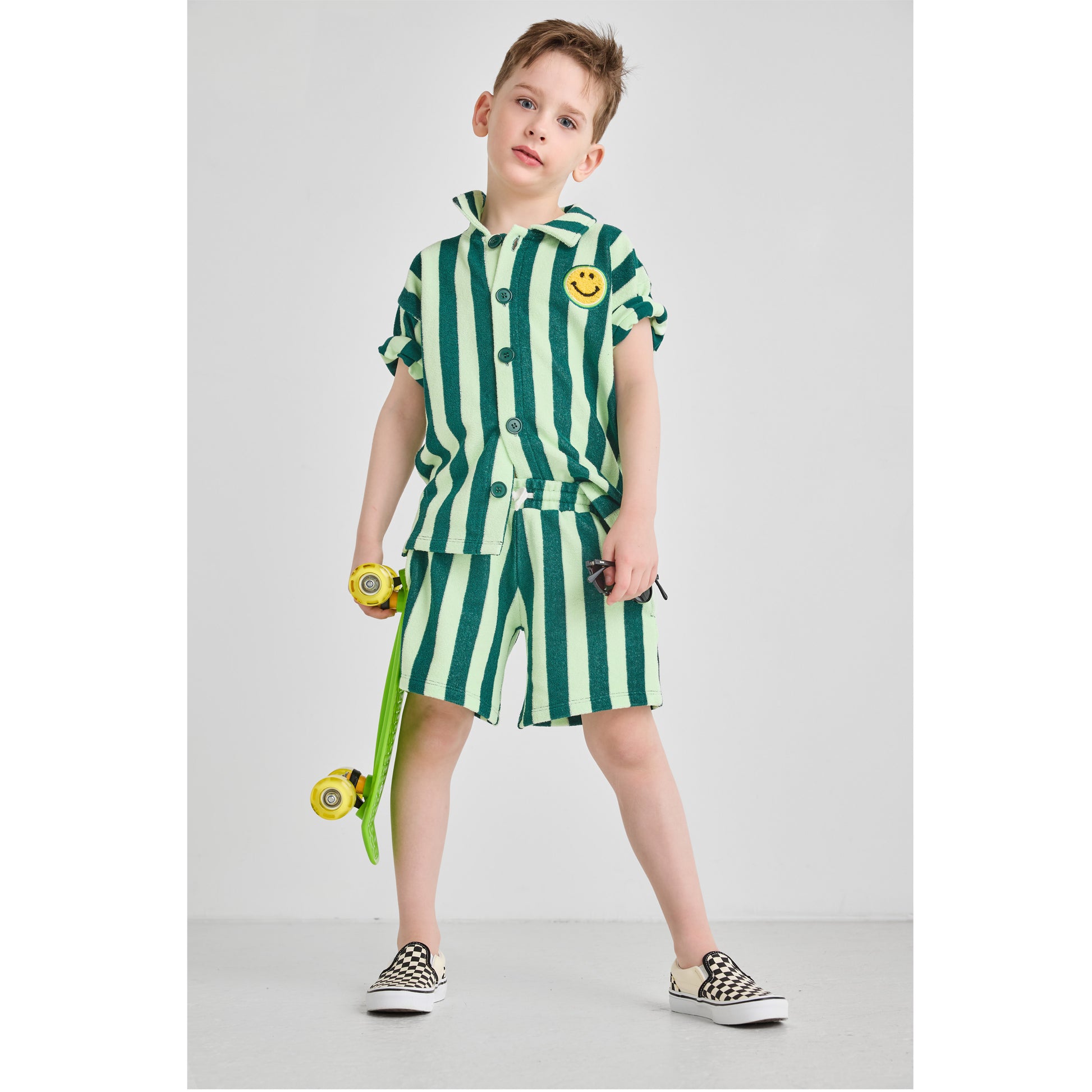 Boy wearing a sustainable, bright green striped set with a smiley face design, holding a skateboard