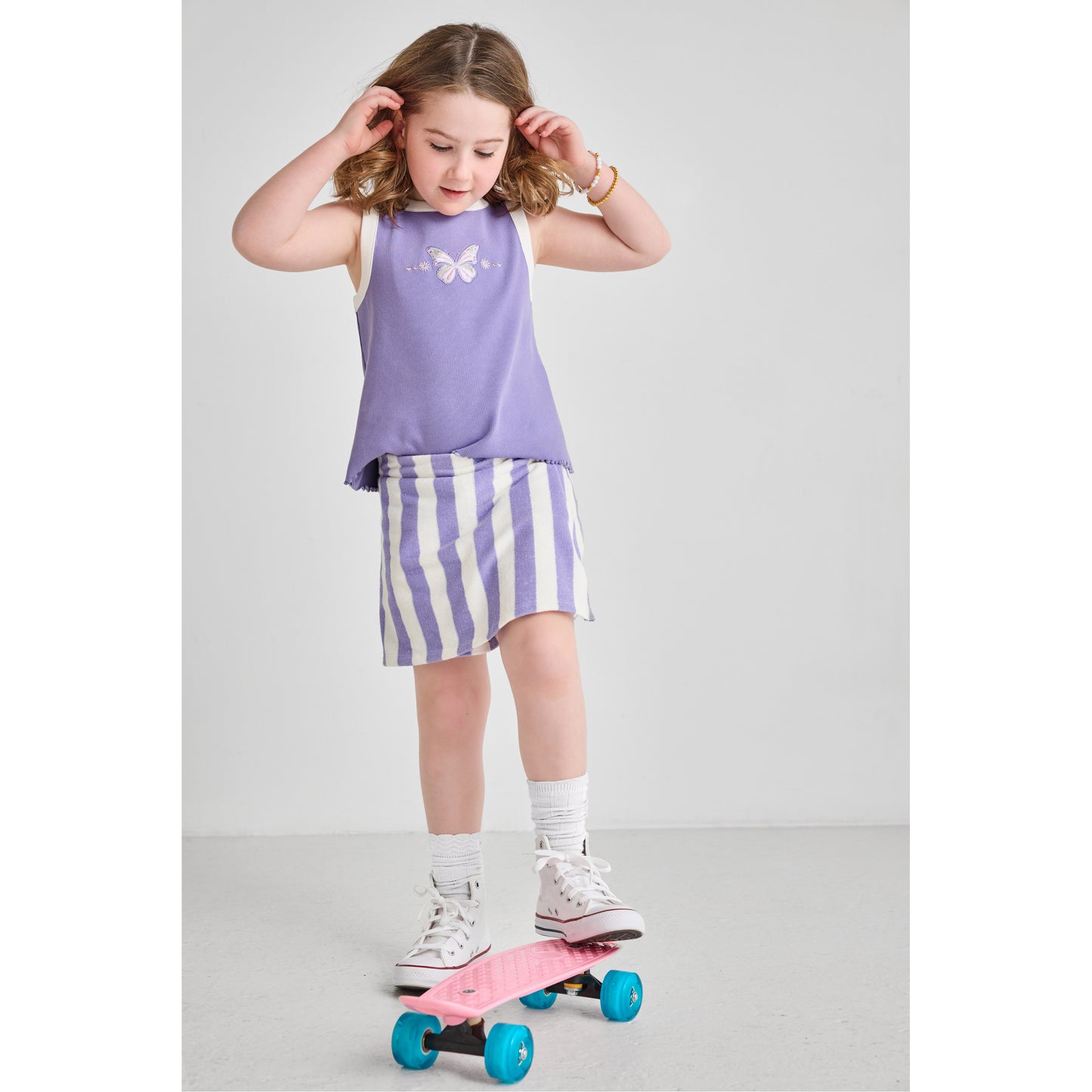 Young girl wearing an eco-friendly organic cotton and recycled polyester purple butterfly tank top and striped skort standing on a pink skateboard