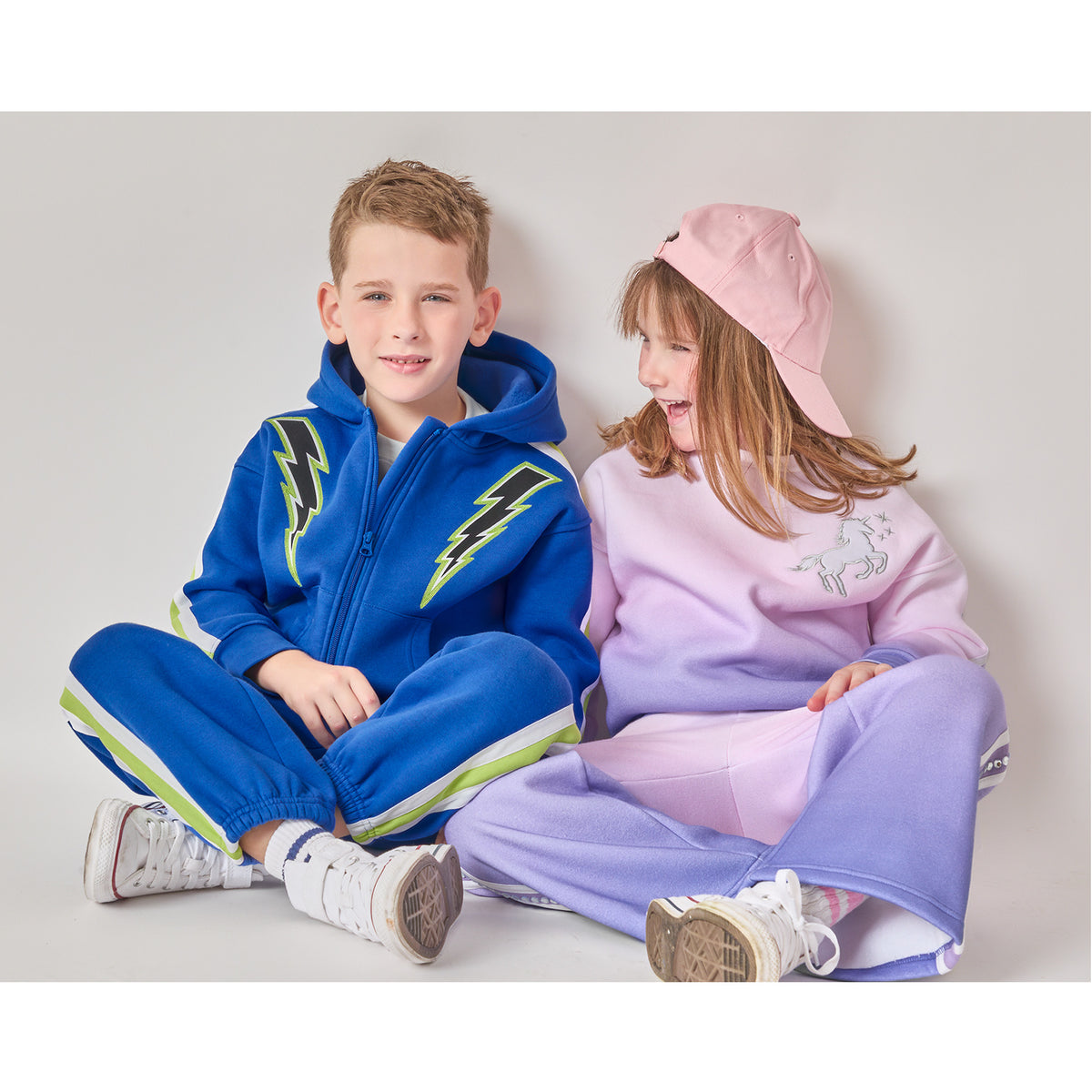 Children sitting side by side in eco-friendly outfits. The boy is wearing a blue lightning zip-up hoodie and matching sweatpants. The girl is wearing a pink-and-purple ombré unicorn sweat set.